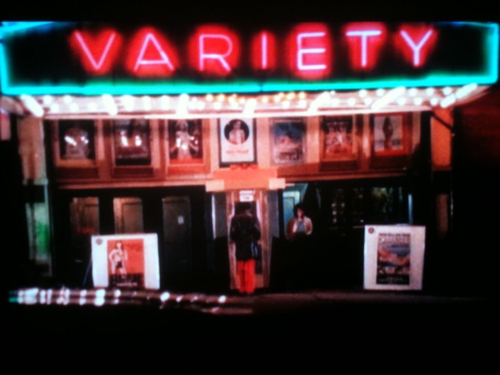 A neon sign that reads 'Variety' over seven framed images and a row of six doors.