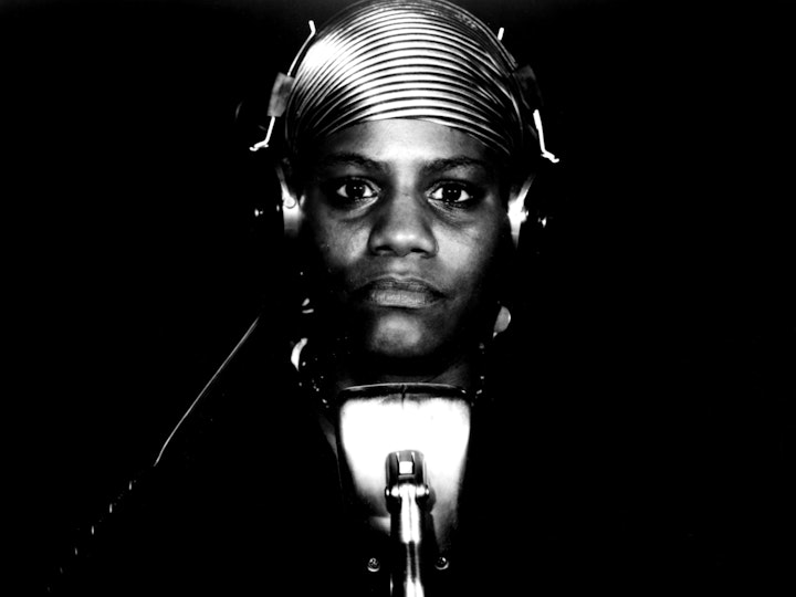 A person with dark skin wears a pair of headphones looks directly into the camera.