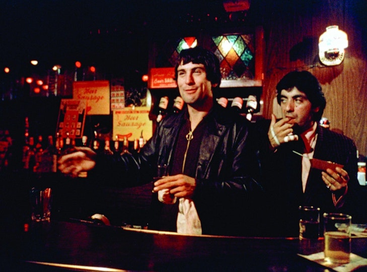 Two people with light skin and black jackets stand behind a bar. One is smiling and the other is licking their finger.
