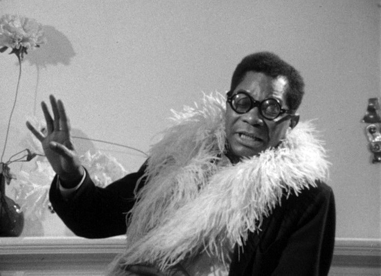 A person with dark skin with round glasses wears a feather boa. Their eyes are closed and one hand is raised.