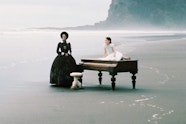 An older woman in a dress stands next to a piano. A younger person in a hite dress sits on top of the piano. The piano is on the shoreline of a beach.