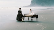 An older woman in a dress stands next to a piano. A younger person in a hite dress sits on top of the piano. The piano is on the shoreline of a beach.