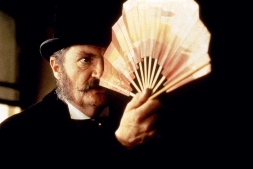 A person with light skin, a beard and moustache in a bowler hat holds a paper fan to their face.