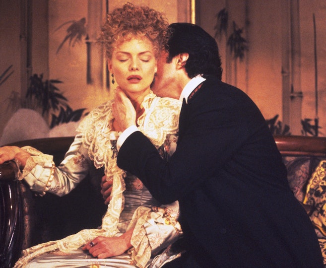 A man kisses the neck of a woman. Both are seated on an ornate sofa.