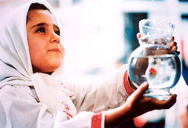 A child with light brown skin in a white headscarf holds a glass vase filled with water.