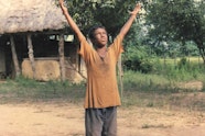 A young person with medium brown skin and dusty clothing raises their hands to the sky. Behind them is a dwelling with a thatched roof and trees.