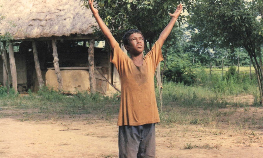 A young person with medium brown skin and dusty clothing raises their hands to the sky. Behind them is a dwelling with a thatched roof and trees.