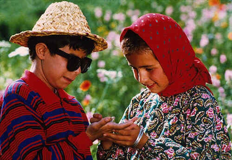 Two children wearing colourful colothes are smiling. One child is holding and looking at the other child's palm. Behind them are flowers. 