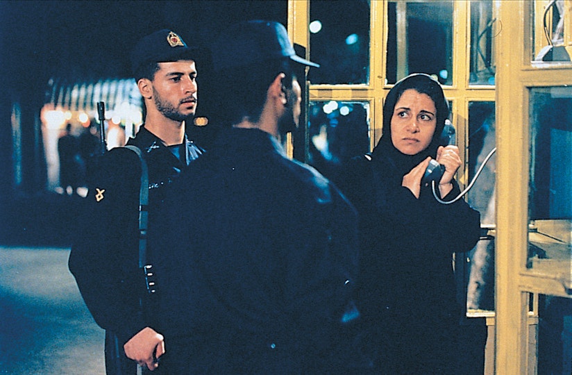 A woman wearing a headscarf using a phone booth. Her hand covers the speaker as two police officers stand closely to her.