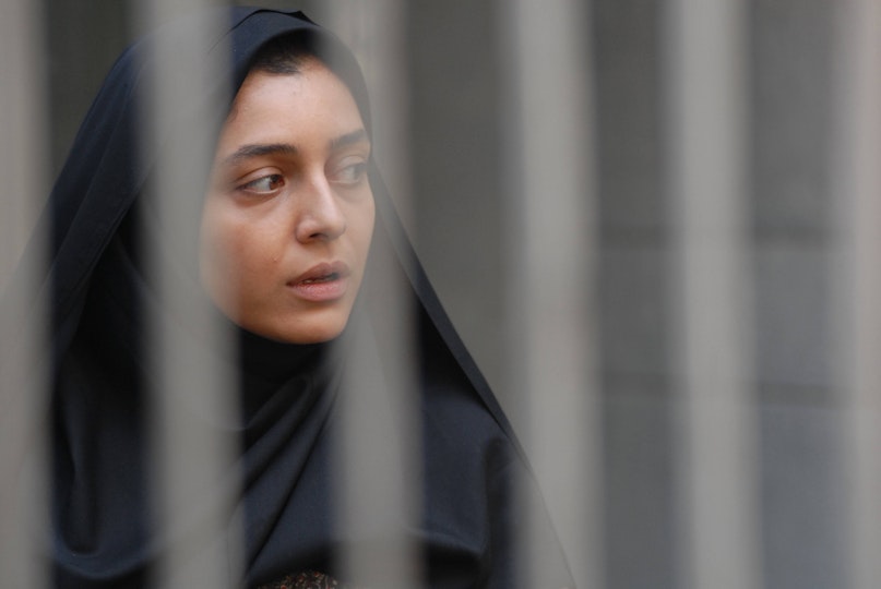 A person with light brown skin and a black headscarf looks over their shoulder. In front of them is a fence.