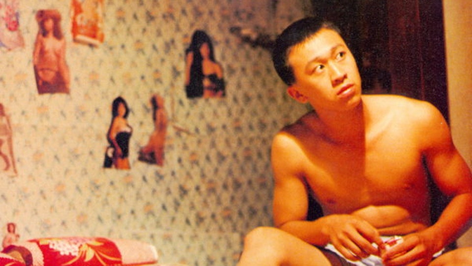 A man with light skin and close-cropped black hair wearing shorts is seated. Behind him are magazine cut-outs of skimpily dressed women glued onto the wall.
