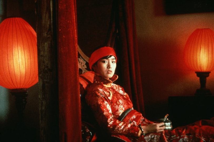 A seated person wearing a red beret and a red and gold silk top looks direcly into the camera. On either side of them are two red lanterns.