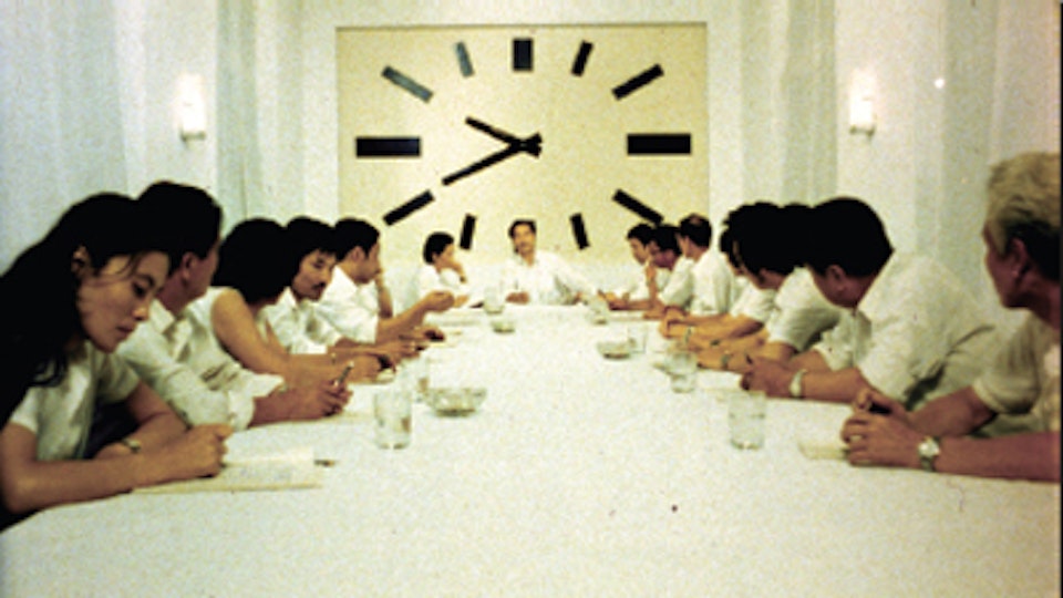 A group of people all wearing white shirts are seated around a long white table. Behind the head of the table is a clock that covers the entire wall.