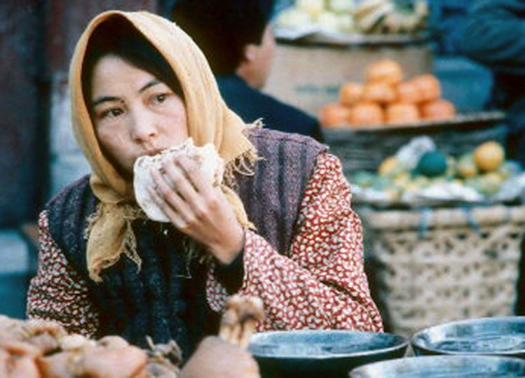 A woman with black hair and a headscarf bites into a sandwich. Behind her is a pile of fruit on top of a basket.
