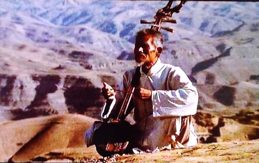 An older person playing a stringed instrument is seated on the ground. Behind them are barefaced mountains.