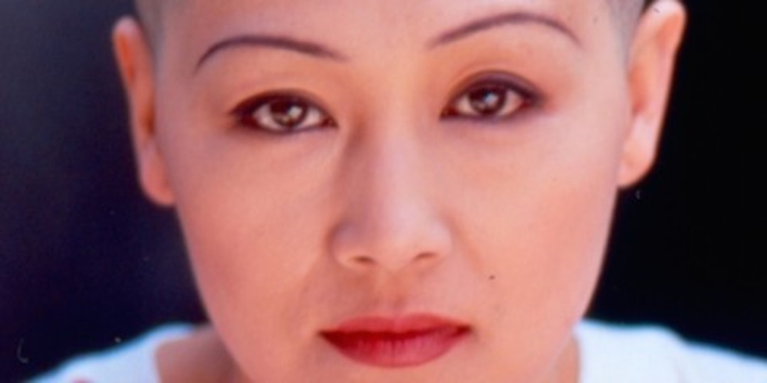A close up of a person with a shaved head, thin eyebrows, brown eyes and bright red lipstick looks into the camera.