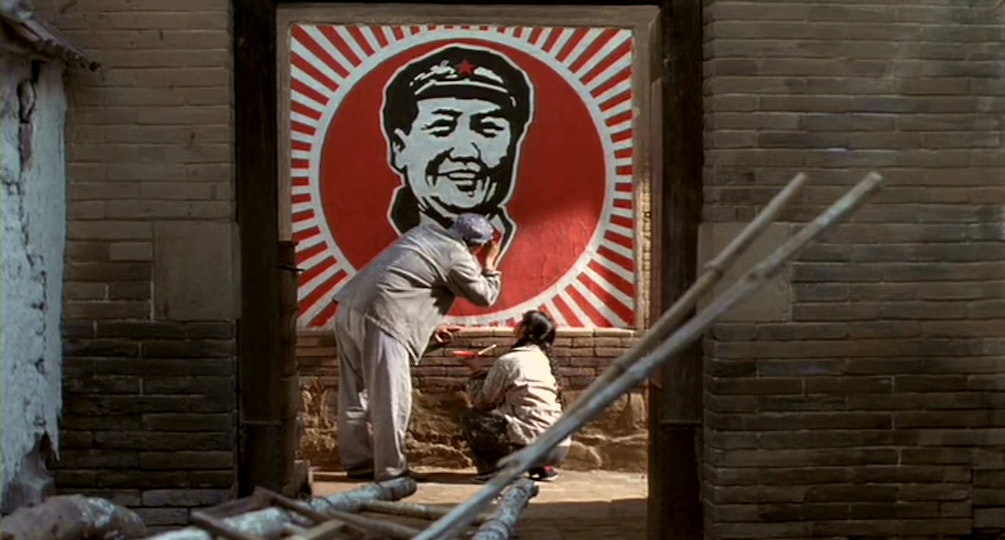Two people in workers clothing stand closely to a brick wall. On the wall is a large illustrated poster of Mao Zedong.