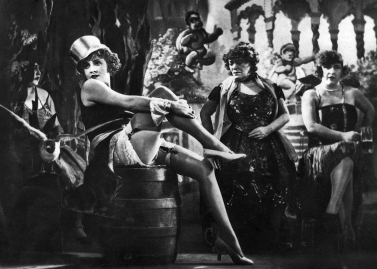 A woman wearing a top hat, dress and high heels sits on a barrel on a stage. Behind her are other women in dresses.