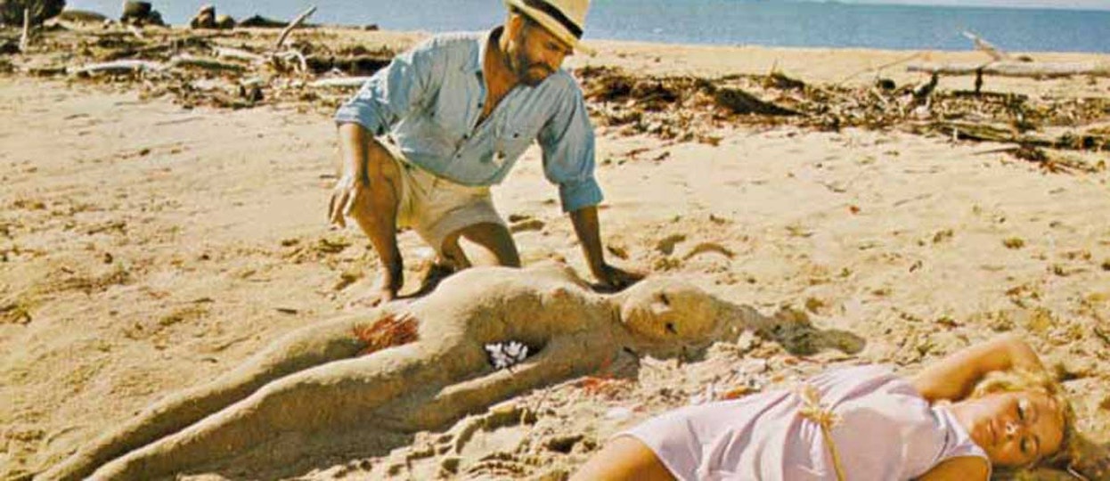 A man on the beach sculpts a figure of a woman lying on the sand. Next to the sand figure is an actual woman lying on the sand.