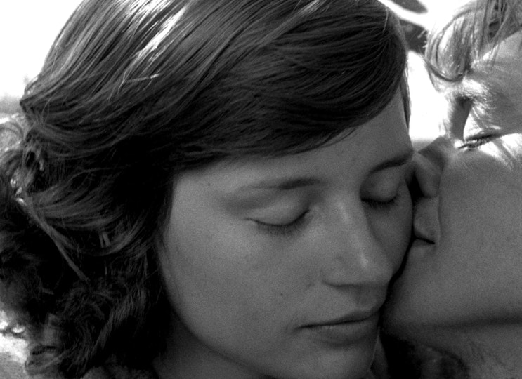 A close up of two women's faces. One woman gently kisses the cheek of the other.