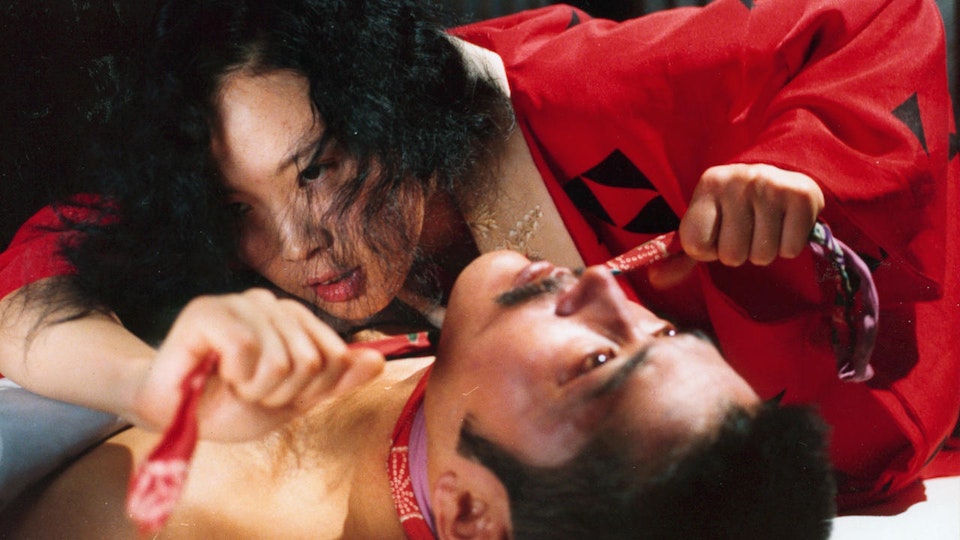 A woman in a red rob ties a scarf around the neck of a prone man.