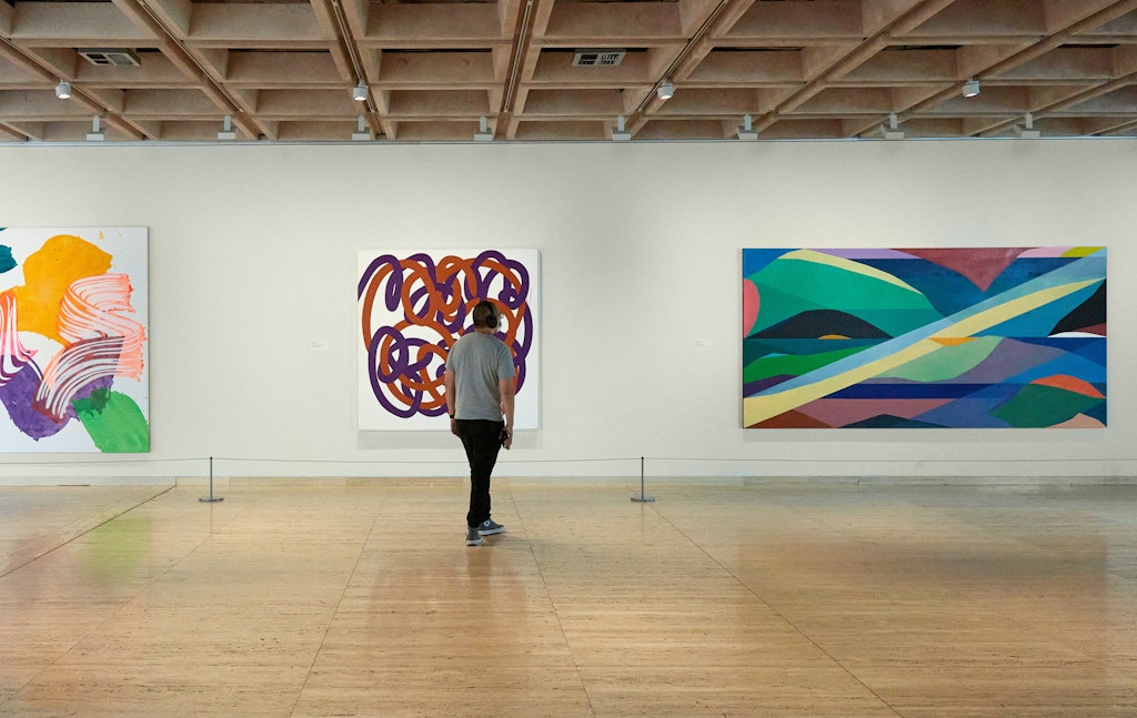 A person stands in front of three colour abstract paintings hanging on a gallery wall.