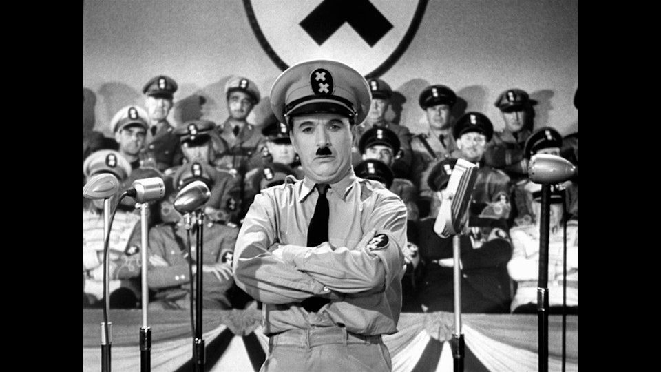 A man impersonating Adolf Hitler stands in front of several microphones and rows of people dressed as Nazi soldiers.