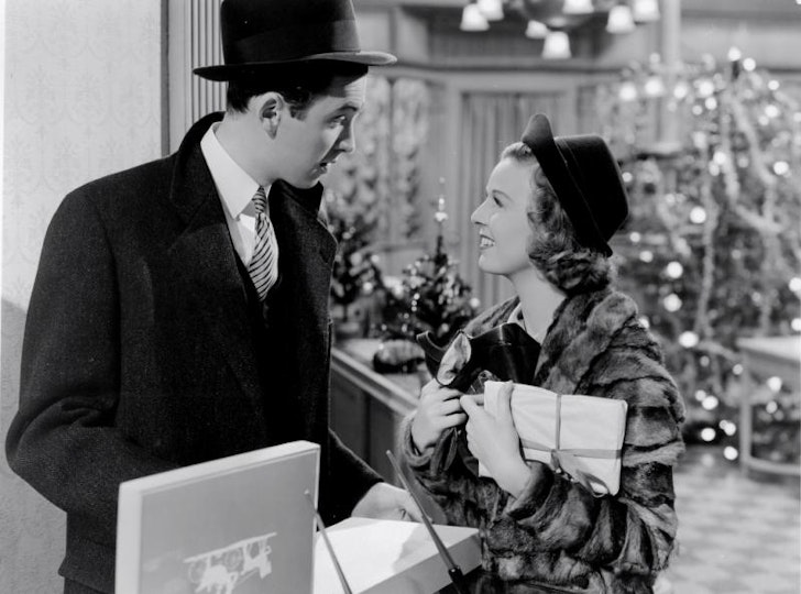Two people wearing coats and hats in a large lobby. The woman on the right is holding wrapped packages.