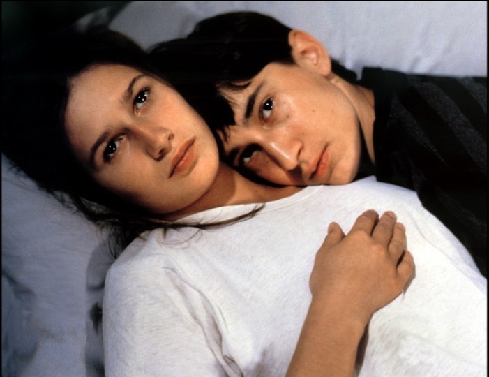 Two people lying together on a bed. One person rests their head on the other's shoulder.