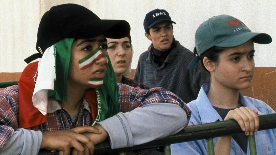Four young people with light brown skin peering over a fence. One of them has painted the green, white and red stripes of the Iranian flag over their nose and chin.