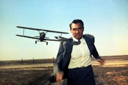 A man with light skin in a suit sprinting towards the camera. Behind him is an airplane flying low to the ground.