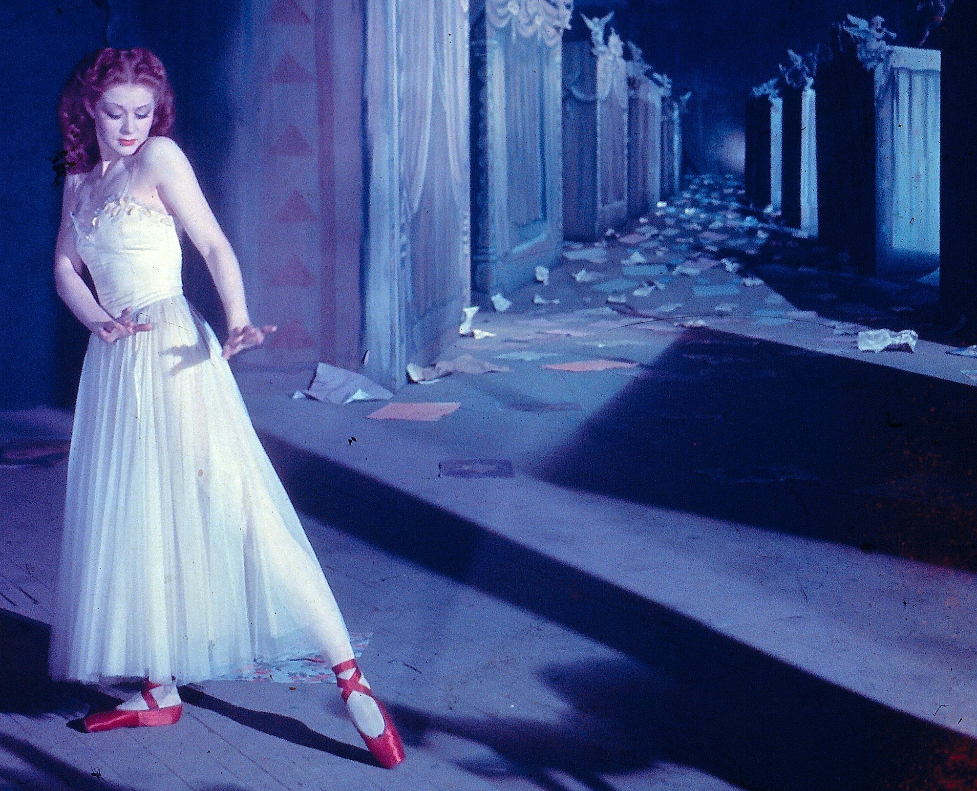 A light skinned woman in a ballet dress and red pointe shoes dances in front of a set strewn with pieces of paper and shadows.