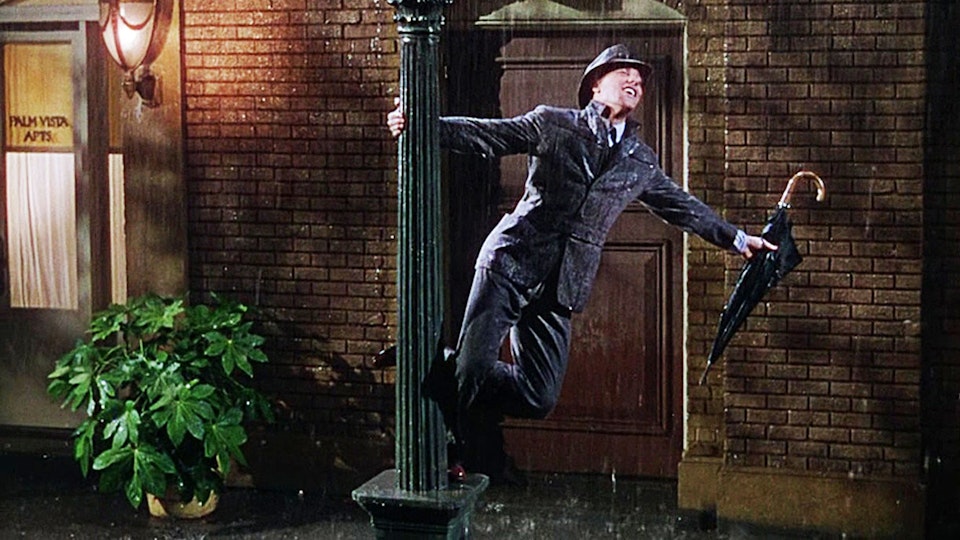 A man swinging around a lamp post in the rain with an umbrella in his hand.