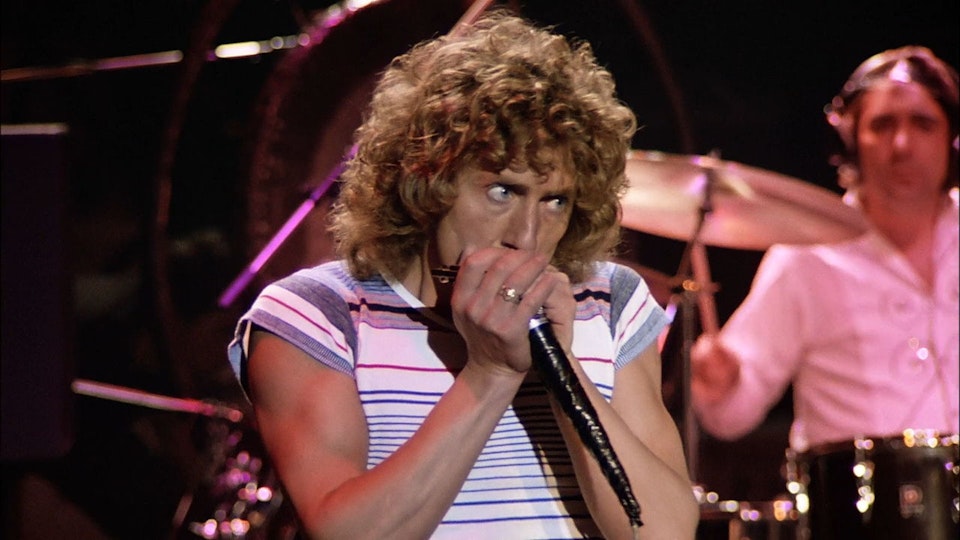 A man with long curly hair and light skin cups a microphone with both hands closely to his mouth.