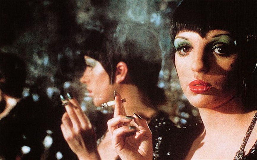 A woman holds a cigarette stands in front of a mirror.