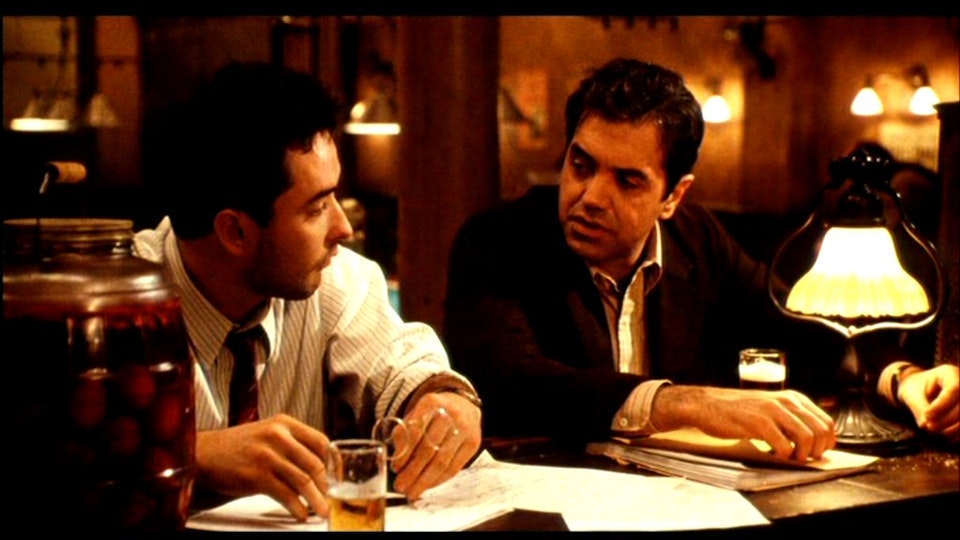 Two men slumping at a bar with beers and paperwork look at each other.