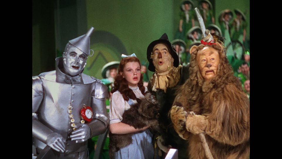 A woman in the middle of a group holding a small dog. Around her is a man a tin costume, a person dressed as a lion, and a person dressed as a scarecrow.