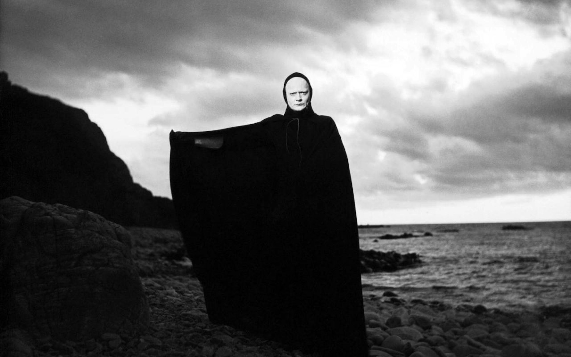 A figure with a white face wearing a floor-length black robe extends an arm. Behind them are cliffs, the sea and clouds.