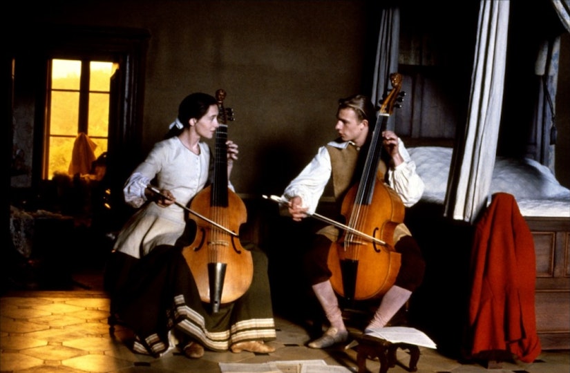 Two people seated in a bedroom playing cellos. They are looking at each other as they play.