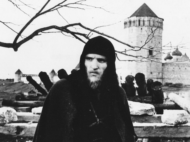 A man with light skin dressed in a dark hooded cape stands in front of fallen fence posts and slabs of stone.
