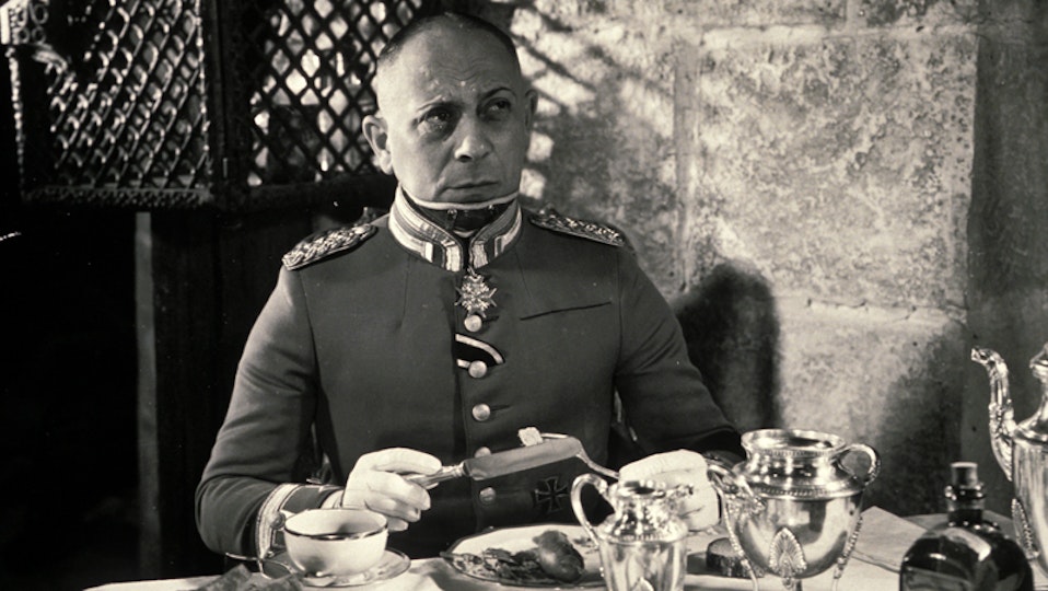 A frowning person in a military uniform is seated at a table. They are holding a knife and fork in their hands.