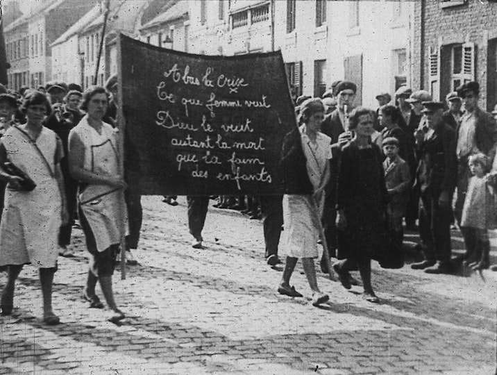 Several women hold a large banner between them. On the banner are cursive lettered words in French. A crowd stares at them.