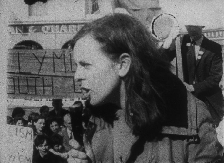 A person with long hair and an open mouth stands high above a crod of protestors.