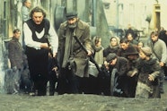 A group of people pulling a cart up a dirt road. Other people in the street are walking behind the cart or looking on.