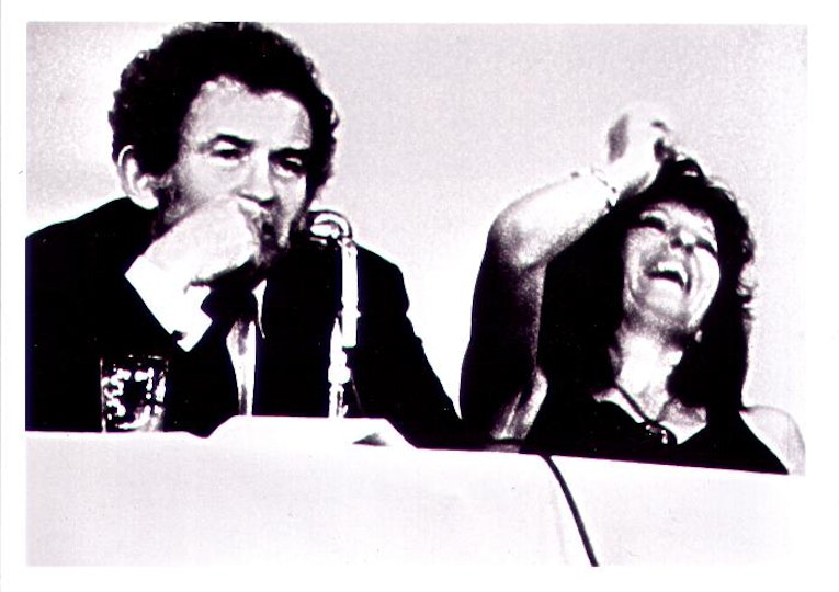 Two people sitting. One person has a microphone in front of them. The other is leaning back and laughing.