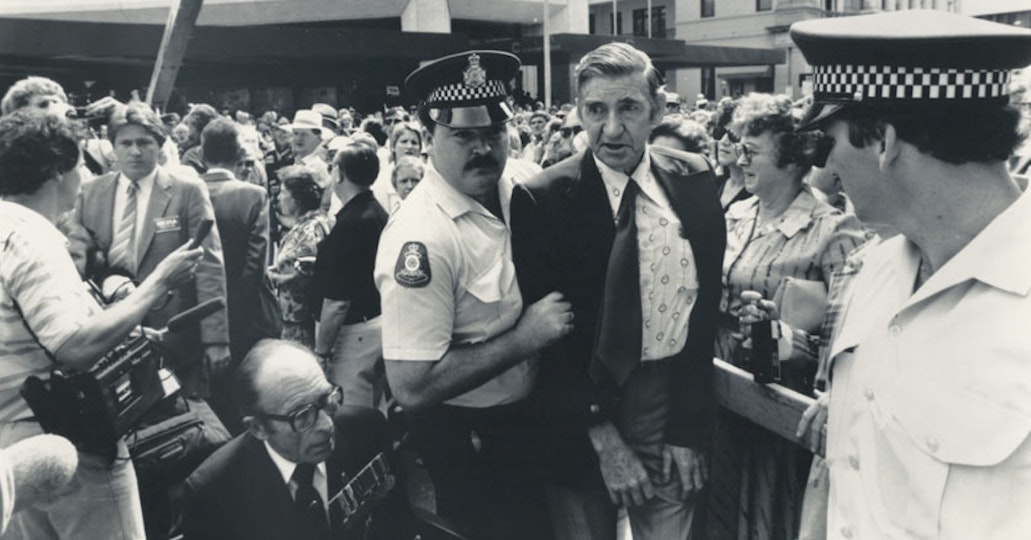 A police officer holds a person in a suit by the elbow. Behind them is a large crowd.