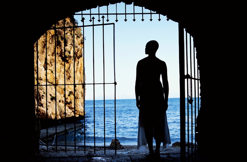 A person, silhouetted in an open gateway, looks out to the ocean and a rocky cliff in front of them.