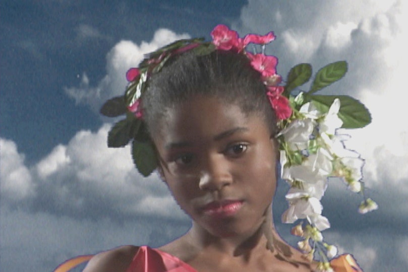 The head and shoulders of a young person with flowers in their hair, against a cloudy sky.