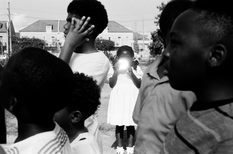 Black-and-white image of a group of young people in the foreground, facing left. Behind them is a young person in a white knee-length, short-sleeved dress, ankle socks and pigtails holding something which flares with light in front of their face. There are buildings and bushes in the background.