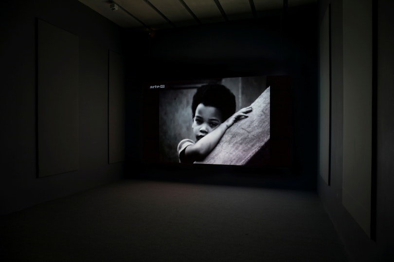Black-and-white image on a screen of a young person with their arm resting on a branch-like diagonal object.